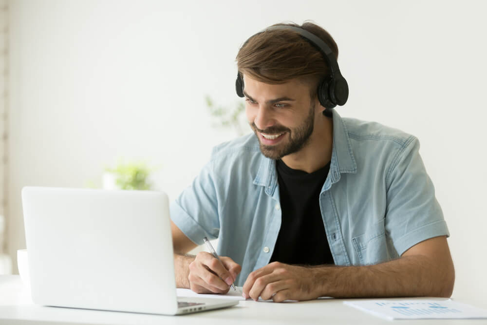 homem animado em frente ao laptop com fones de ouvido e fazendo anotaçoes