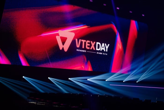 telão do VTEX Day 2026, com etalhes azul e rosa, e embaixo há um jogo de luzes para diversas direções.