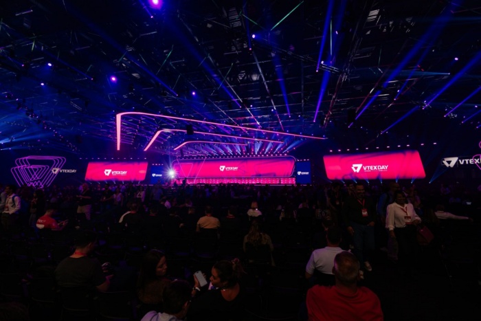  imagem de grande público reunido em frente aos telões do VTEX Day 2026.
