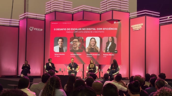 foto do painel de Rafael Mayrink com mais três pessoas, incluindo Graciela Kumruian, da Netshoes, que está segurando o microfone, todos sentados. No fundo há pilares rosas e uma projeção do título da palestra com as imagens dos participantes.