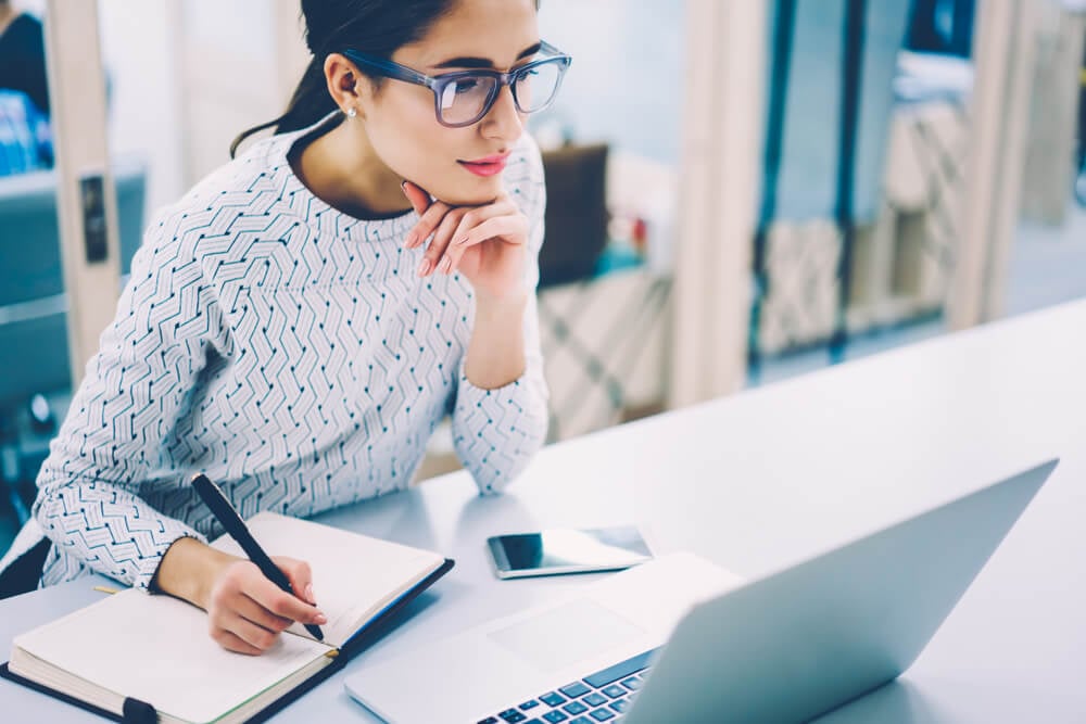 mulher concentrada fazendo anotações e olhando para laptop