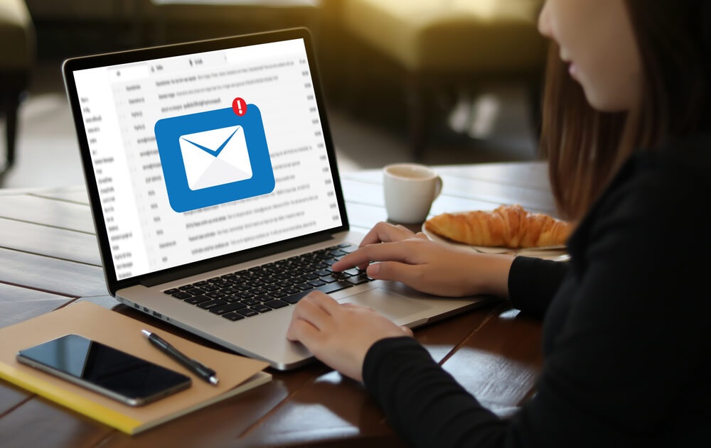 mulher em mesa de escritorio checando emails em laptop com xicara e smartphone ao lado